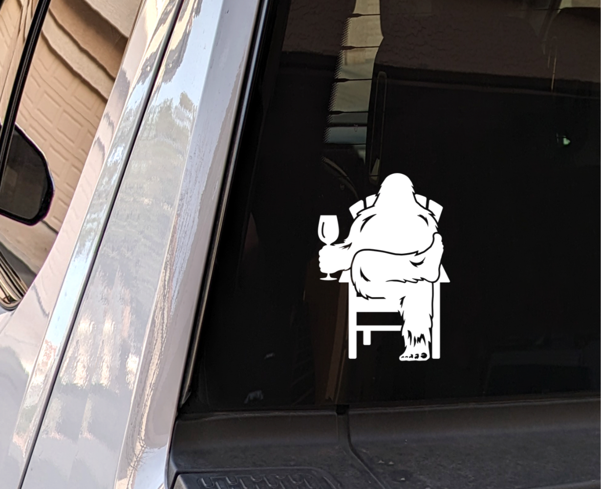 White decal of Bigfoot relaxing in a chair and enjoying a glass of wine, on the rear window of a truck