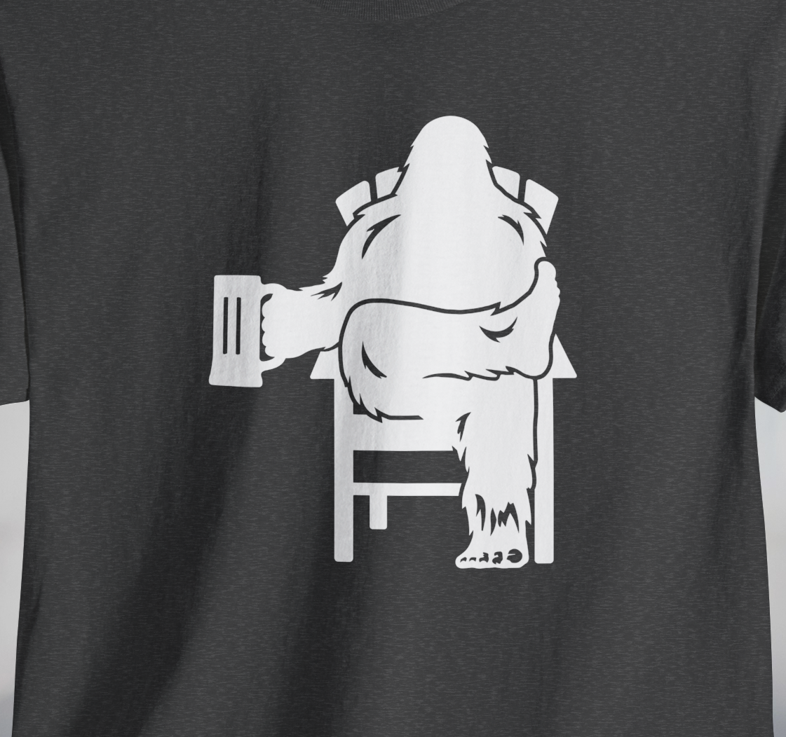 Gray t-shirt with white graphic of Bigfoot holding a beer mug, sitting in a lawn chair with leg crossed in relaxed position.