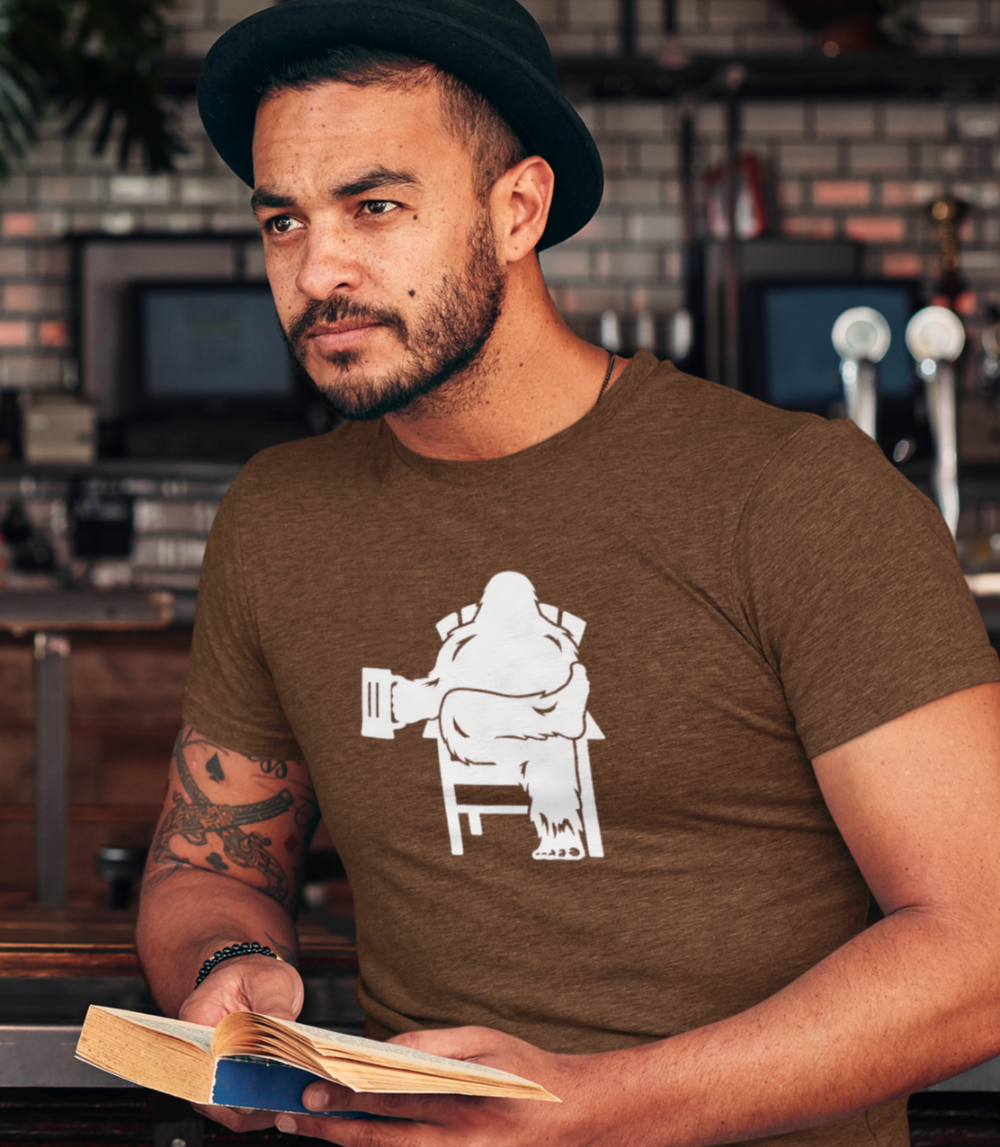 Man wearing a brown shirt with white graphic of Bigfoot relaxing in a chair and enjoying a mug of beer.