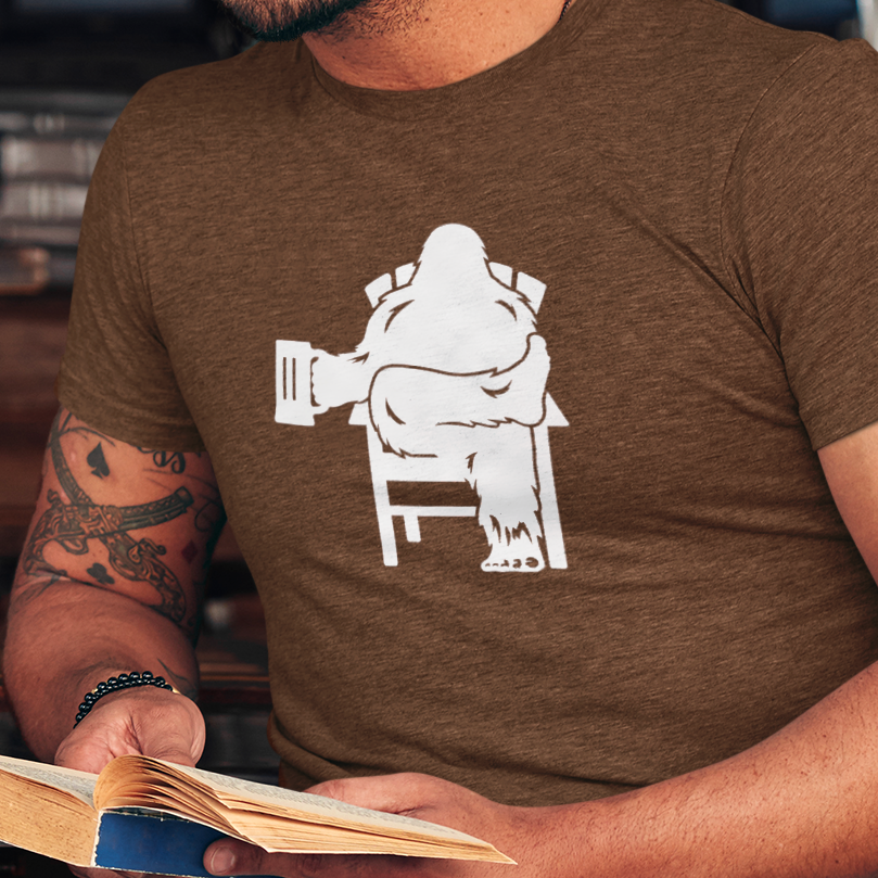 Man wearing a heather brown t-shirt with graphic of Bigfoot holding a beer mug, sitting on a lawn chair relaxing