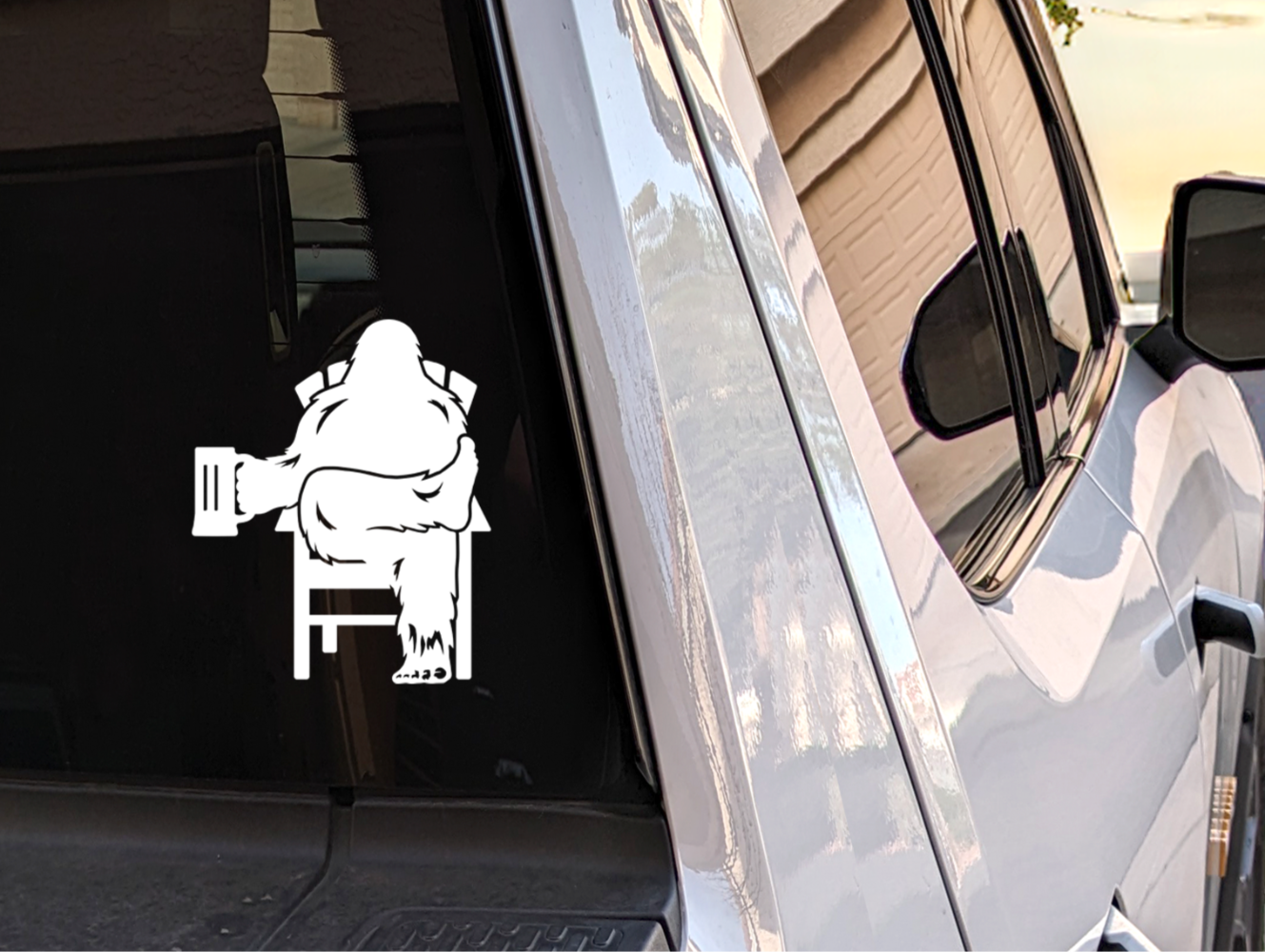 Truck rear window with white decal of Bigfoot relaxing in a chair holding a beer mug.