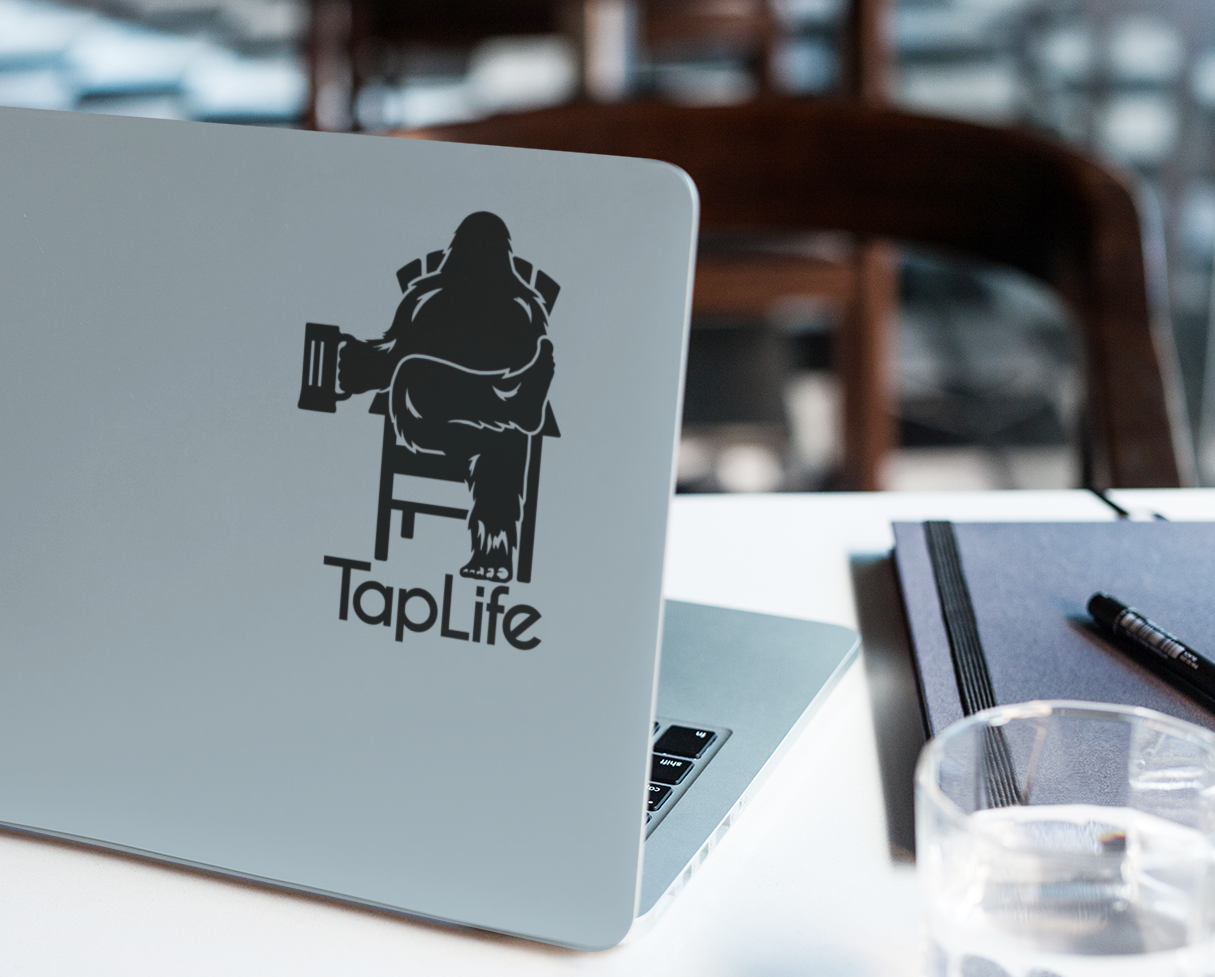 Laptop with black decal of Bigfoot relaxing in a chair, holding a beer mug, with Tap Life logo below.