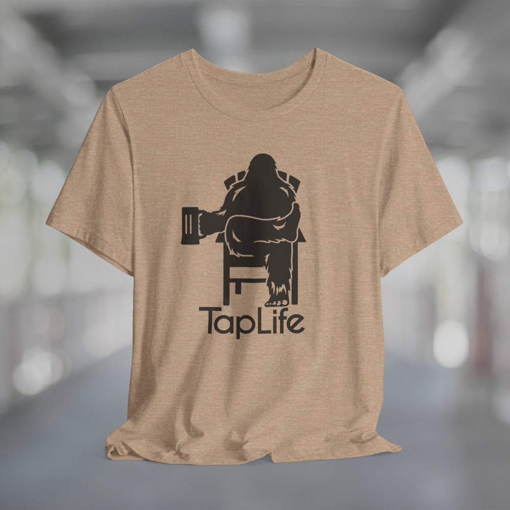 Heather tan t-shirt with black graphic of Bigfoot holding a beer mug, sitting in a lawn chair with leg crossed in relaxed position, Tap Life logo below.