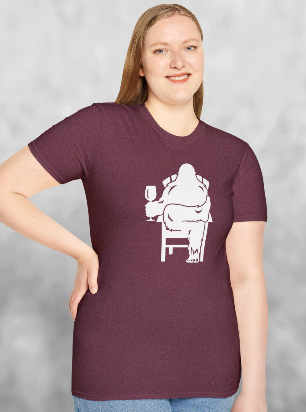 Woman wearing Heather red maroon t-shirt with white print of Bigfoot relaxing in a chair and enjoying a glass of wine.