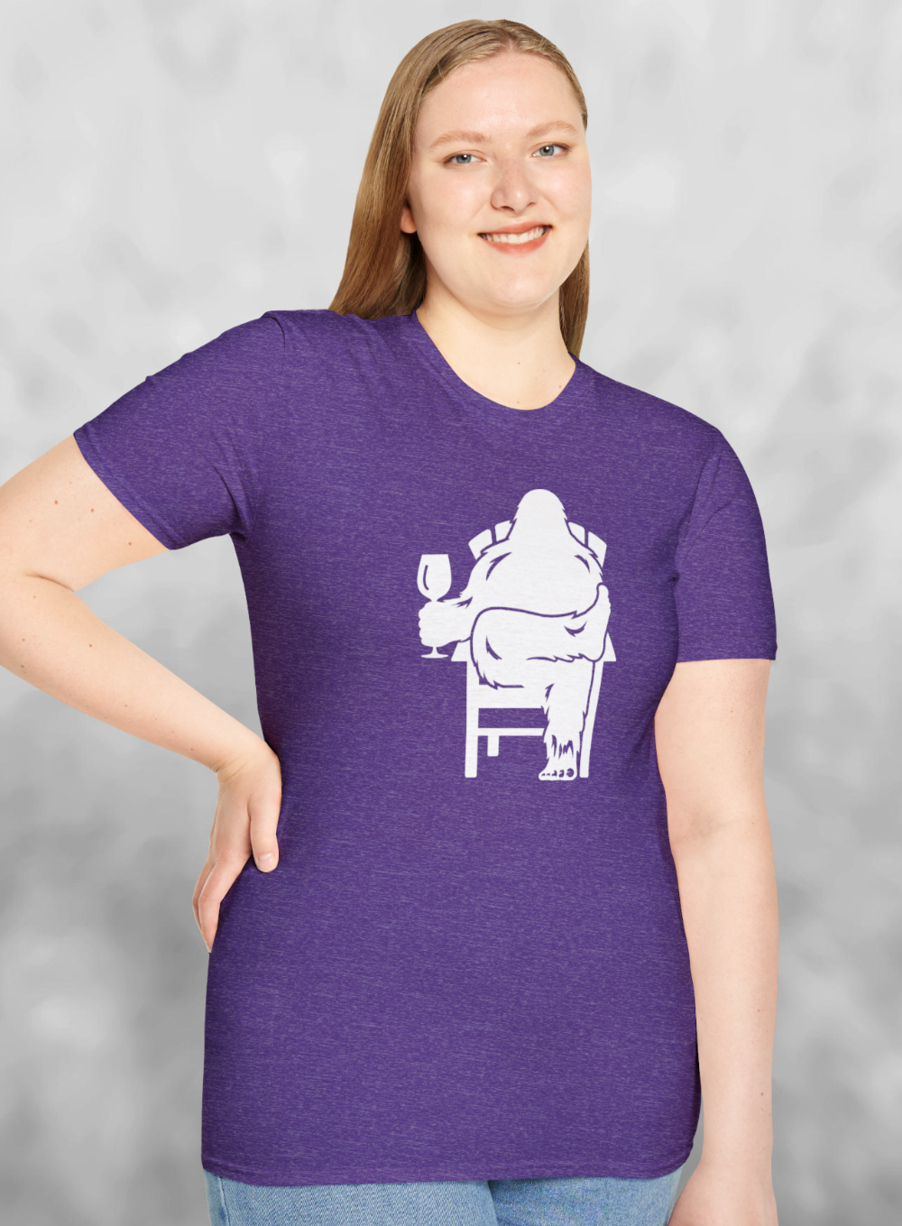 Woman wearing Heather purple t-shirt with white print of Bigfoot relaxing in a chair and enjoying a glass of wine.