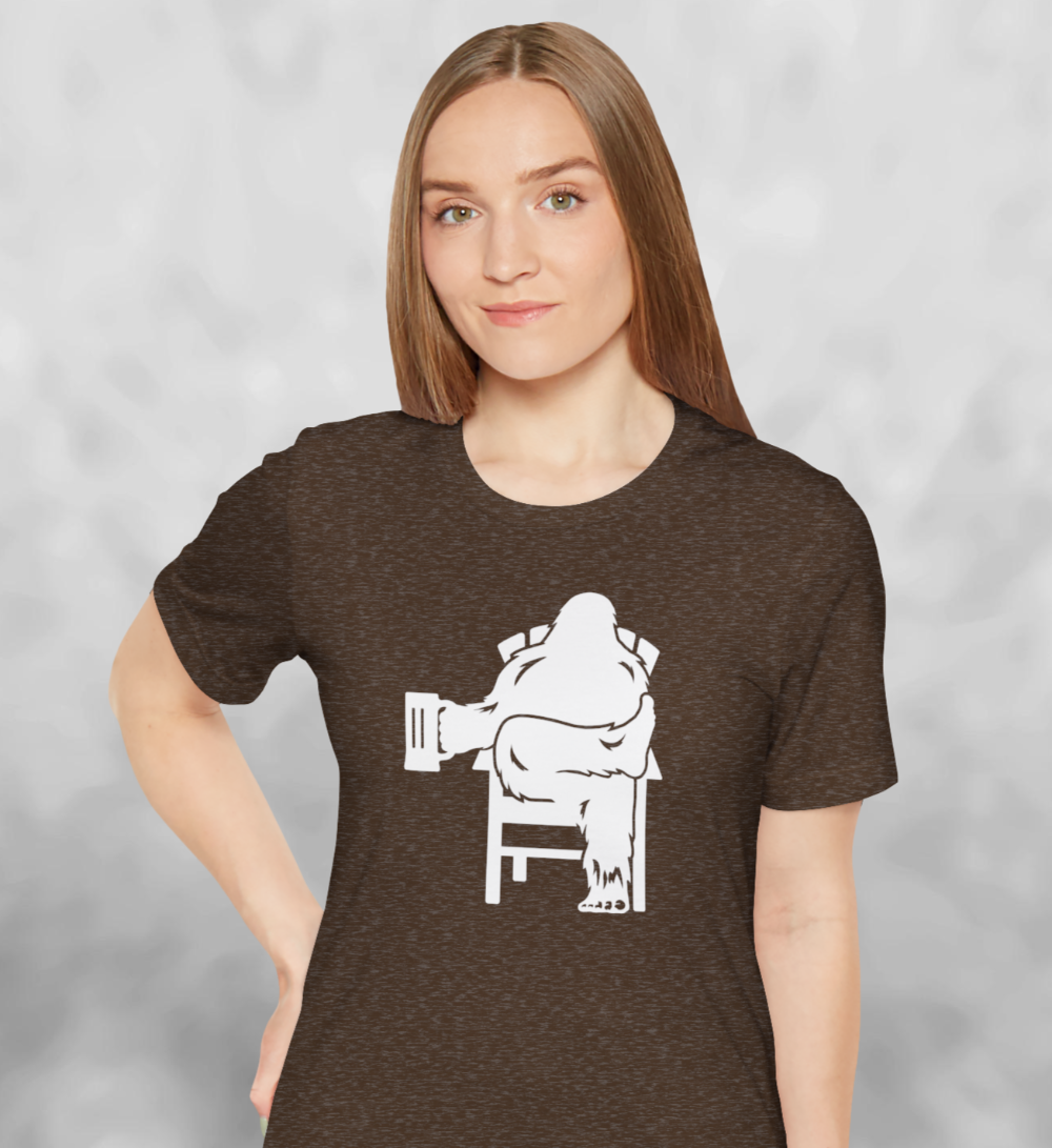 Woman wearing heather brown t-shirt with white graphic of Bigfoot relaxing in a chair and enjoying a mug of beer.