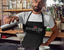 Man mixing a cocktail while wearing a black apron, with a graphic design of the red Wine Life logo across the front.