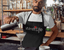 A bartender wearing a Wine Life Apron, pouring a cocktail at a stylish bar.