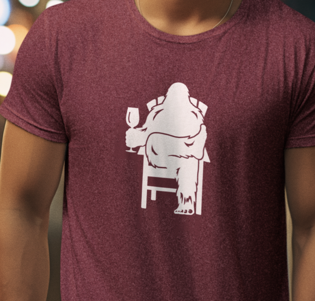 Man wearing a heather maroon shirt with graphic design of Bigfoot holding a wine glass, while sitting in a lawn chair relaxing