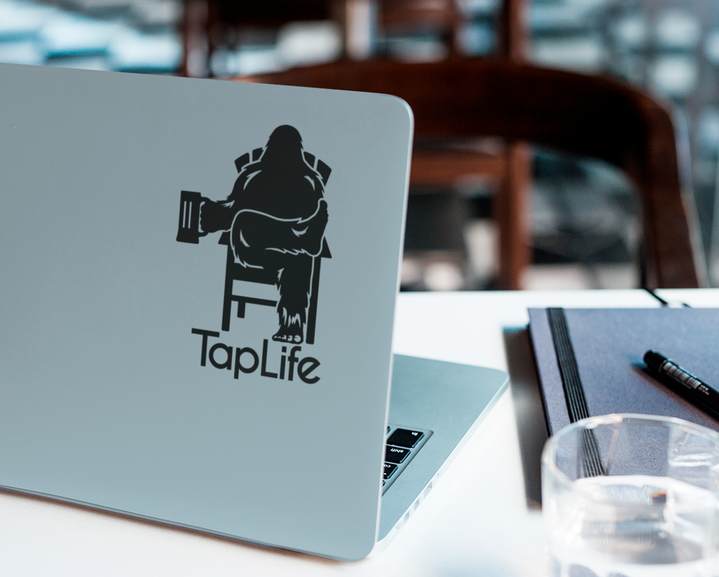 Laptop with black decal of Bigfoot relaxing in a chair, holding a beer mug, with Tap Life logo below.