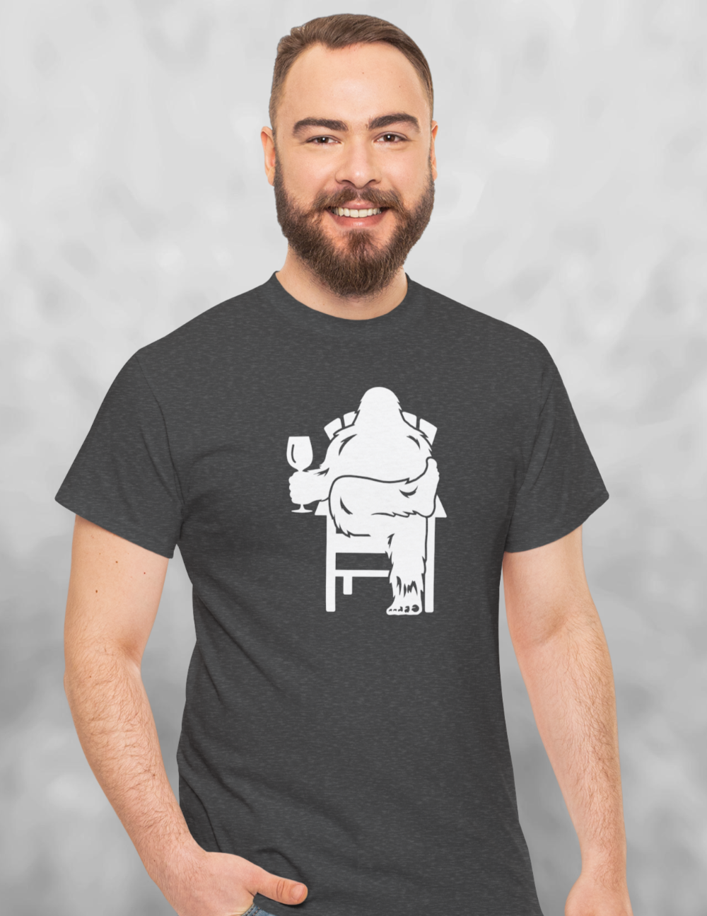 Man wearing Heather charcoal gray t-shirt with white print of Bigfoot relaxing in a chair and enjoying a glass of wine.