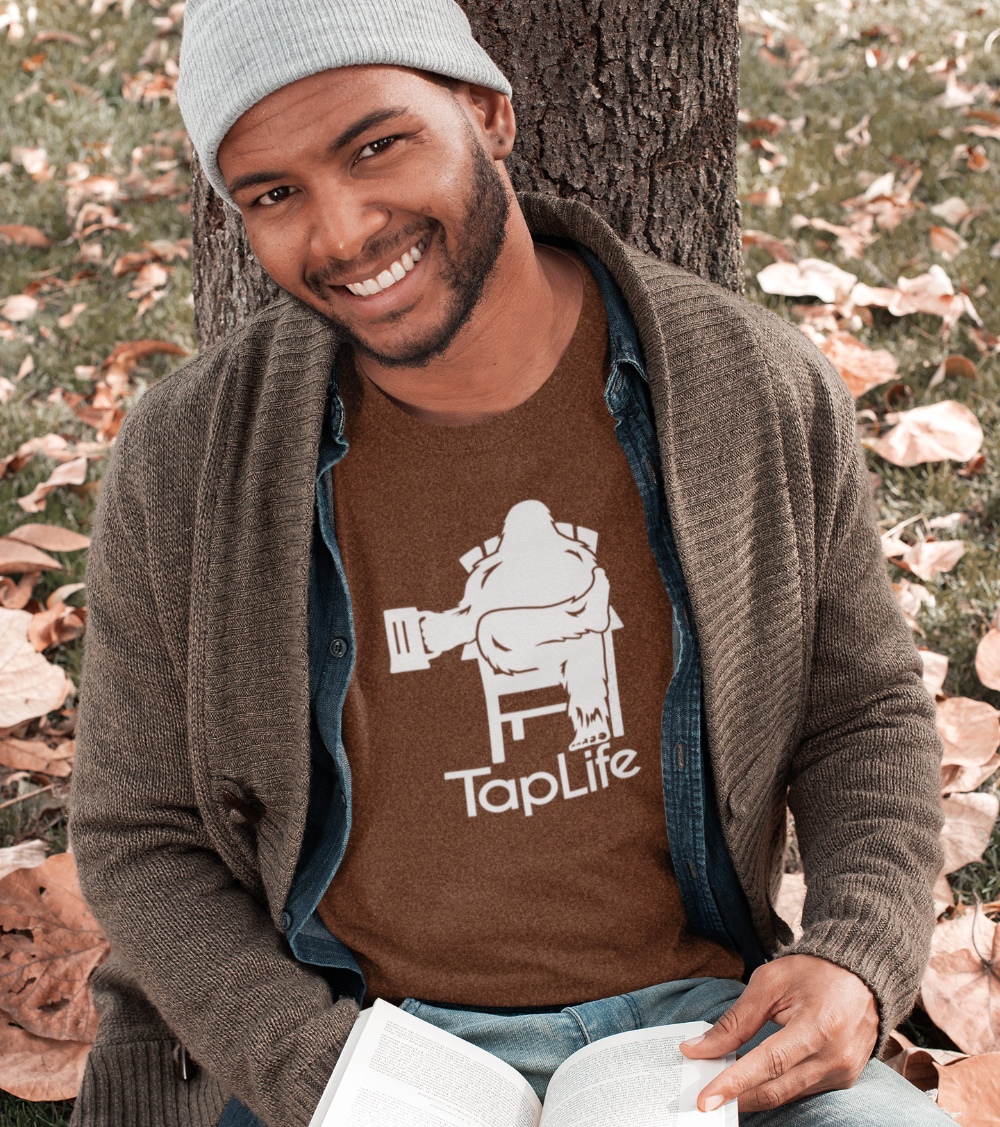 Man wearing Heather brown t-shirt with white graphic of Bigfoot holding a beer mug, sitting in a lawn chair with leg crossed in relaxed position, Tap Life logo below. Main image